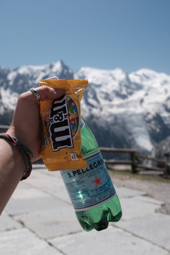 A hand holding M&M candies and a drink against a backdrop of mountains that are out of focus.